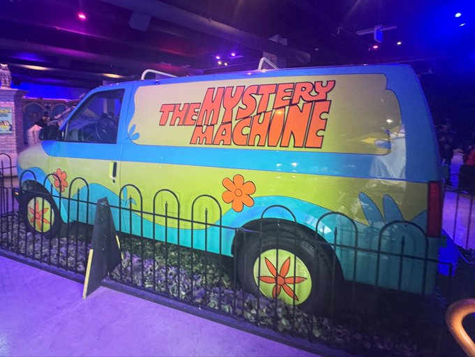 A replica of the Scooby-Doo 'Mystery Machine' van displayed indoors behind a small black fence, illuminated with purple lighting.