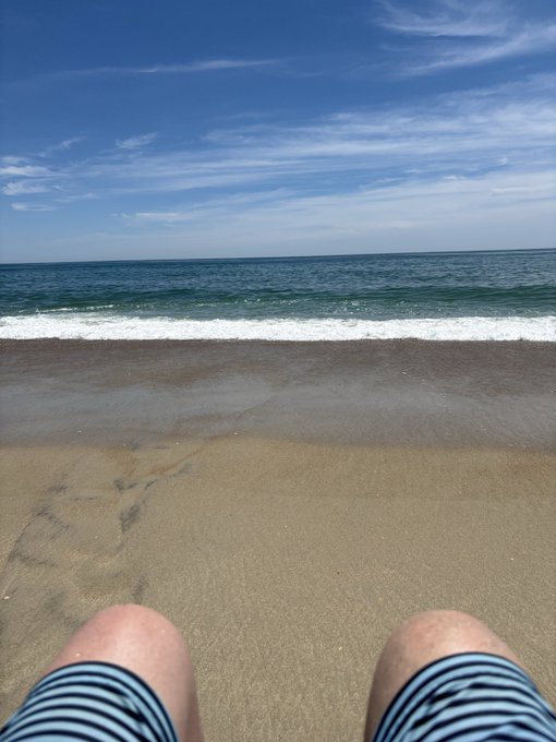The image shows a person's legs with their feet visible, wearing striped swim trunks. The beach setting features a sandy shoreline with waves crashing onto the shore. In the background, there are clouds, suggesting that the weather might be overcast or changing. The photo appears to have been taken from someone's perspective on the beach, capturing both the person and the surrounding environment.