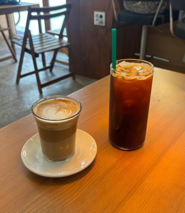 A small espresso/cortado in a glass cup next to a tall iced coffee with a green Starbucks straw sit on a wooden table in a cafe.
