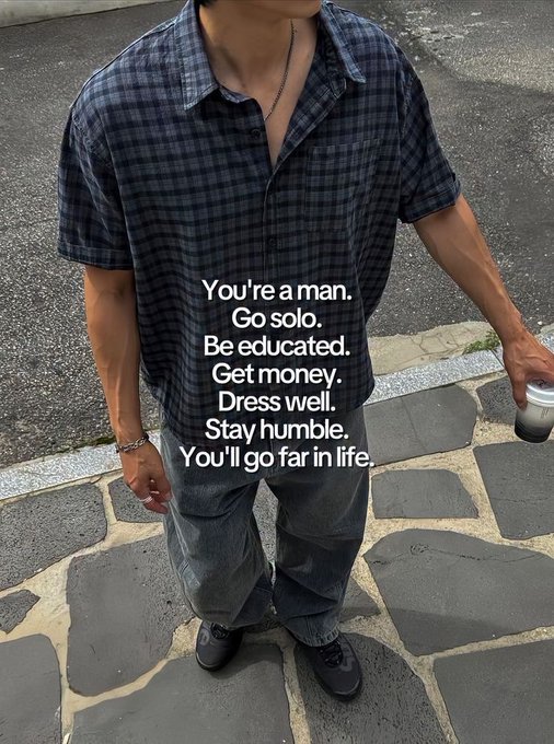 The image features a man standing on a sidewalk, wearing a plaid shirt and jeans. He has his arm outstretched as if he's about to say something or make a gesture. The background of the image shows a street with a parking lot visible behind him. There is also a cup placed near the edge of the frame.