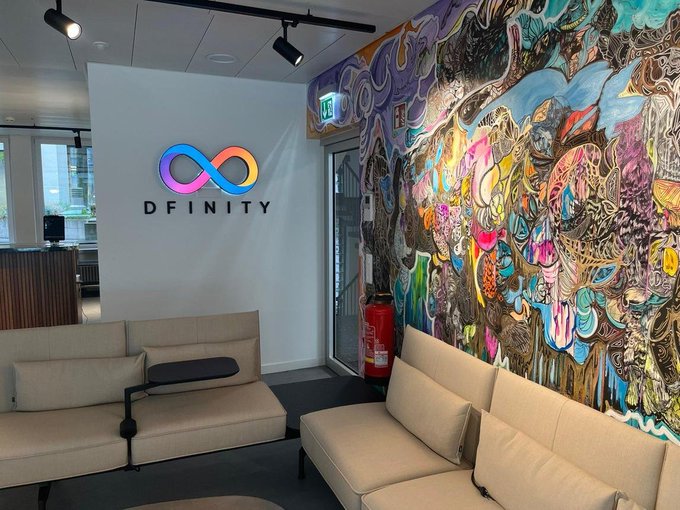 A modern lounge area featuring a wall with a colorful infinity logo and the text 'DFINITY', alongside a vibrant abstract mural and beige seating.