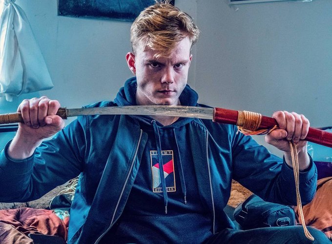 A young man with blonde hair wearing a blue hoodie, sitting on a bed and holding a large knife or machete with a red handle across his neck, staring intensely at the camera.