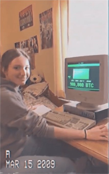 A woman sitting at an old desktop computer with a monitor displaying a wallet showing "500,000 BTC", dated "MAR 15 2009".