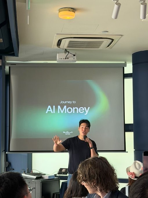 A man speaks into a microphone on a stage in front of a projection screen displaying 'Journey to AI Money,' with an audience seated in the foreground.