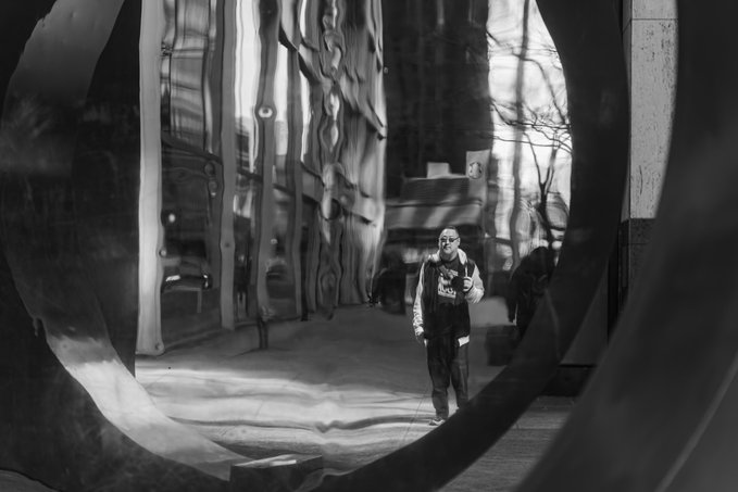 A black-and-white photograph captures a man wearing sunglasses standing in the distance, viewed through the circular reflection of a dark, metallic sculpture in an urban setting.