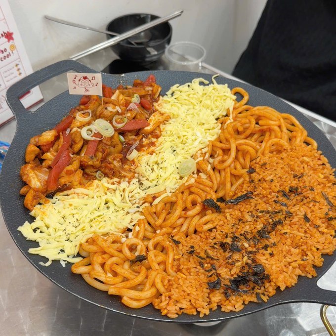 A top-down photograph of a Korean dish served in a black pan, containing stir-fried chicken and red peppers, melted cheese, spicy noodles, and seasoned rice topped with seaweed.