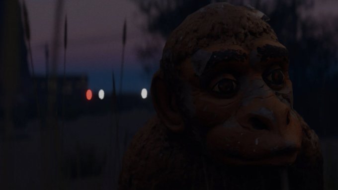 A dark, moody film still showing a close-up of a monkey or ape statue/figure with a tear on its face, set against a twilight background with blurred lights.