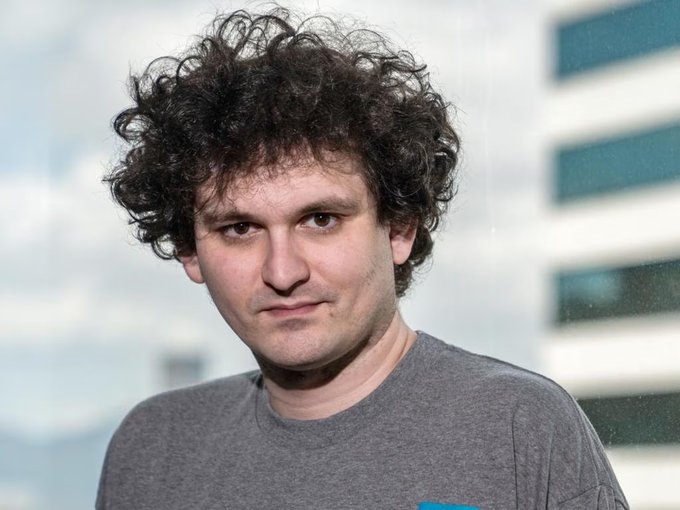 A man with curly dark hair wearing a gray t-shirt looks directly at the camera, with a blurred office building visible through the window behind him. The image relates to the post as it appears to show Sam Bankman-Fried, the FTX founder whose clemency bid was rejected by the U.S. Senate.