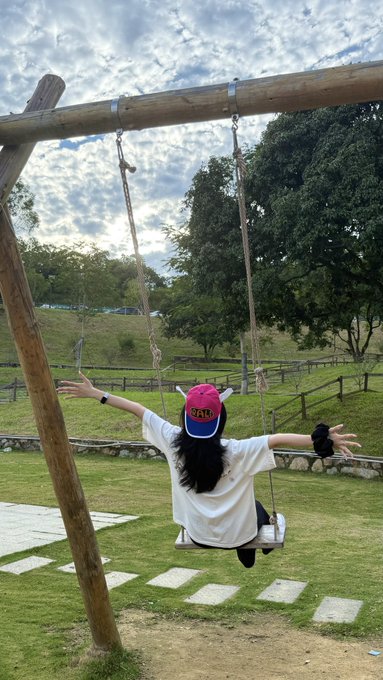 A young woman with long dark hair, wearing a white shirt and pink-brimmed cap, sits on a wooden swing with her arms outstretched against a backdrop of green fields and trees under a cloudy sky.