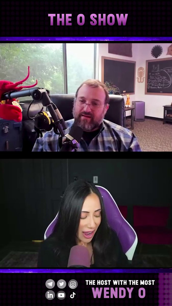 The image shows a man and woman sitting at a table, with one of them holding a microphone while they appear to be having an interview or discussing something related to podcasts. The other person seems to be listening intently as well. There are two microphones in the scene - one being held by the person on the left side of the image, and another one placed near the center-right area.

The background features a window with blinds that can be seen partially through it. A chair is also visible in the room, positioned close to the window. The overall setting seems to be an indoor space where people gather for discussions or interviews related to podcasts or other audio content.