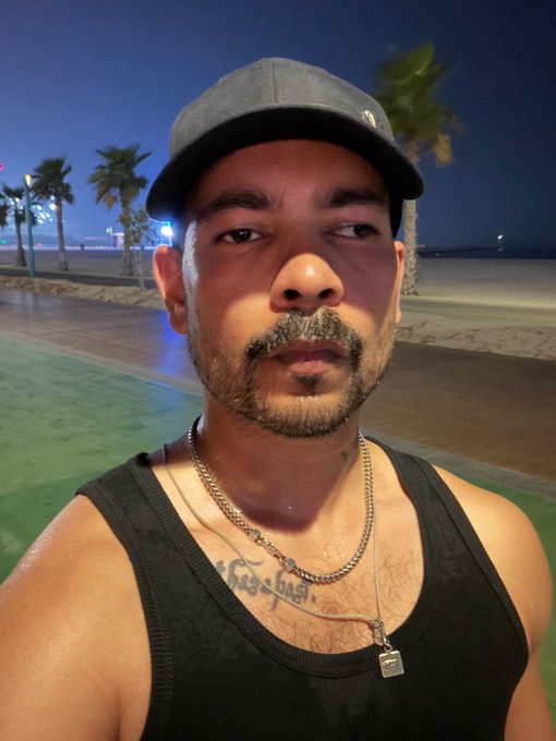 A man wearing a black tank top, cap, and silver chain poses for a selfie at night along a palm-lined beachfront promenade, with a tattoo visible on his chest. The image complements the post's fitness-focused message about completing a workout and evening cardio on Tuesday, embodying a routine that goes well beyond average effort.
