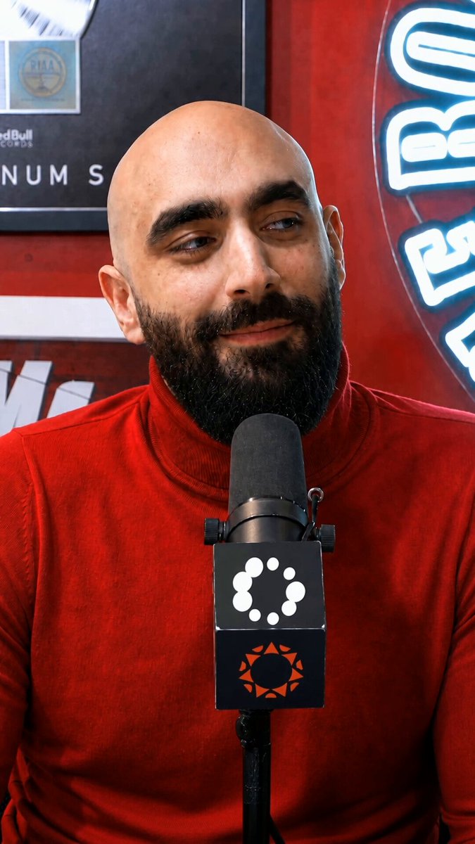 The image features a bald man wearing a red shirt, sitting behind a microphone with his head tilted back as if he's speaking into it. The background includes a poster of a beer sign and a picture of a person holding a beer can. There is also an orange circle on the bottom right corner of the image. The post reads "Turnt it down just to grind for Solana.", which seems to be related to the man speaking into the microphone or possibly discussing the concept of grinding in the context of a podcast or interview.