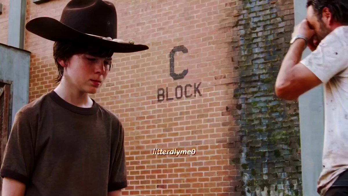 The image shows two people standing next to each other against a brick wall with the word "BLOCK" written on it. The person on the left has a cowboy hat, while the person on the right wears a white shirt. They appear to be engaged in conversation or observing something together.