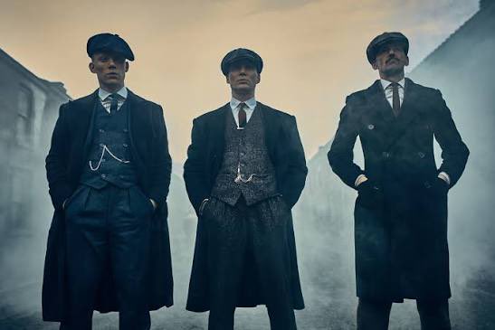Three men in dark overcoats and flat caps stand in a smoky street, evoking the gritty aesthetic of the TV series *Peaky Blinders*, with the atmospheric imagery likely chosen to symbolize the sharp, power-driven themes of macroeconomics and markets discussed in the post.