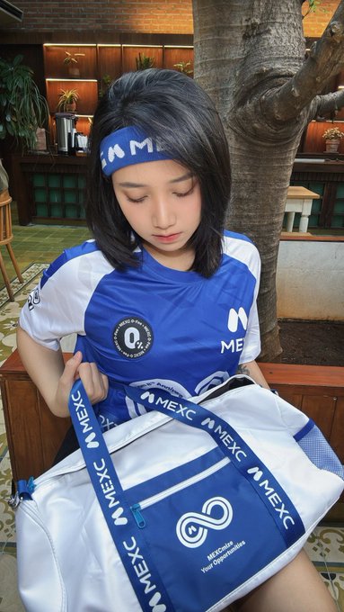 A woman wearing a blue and white MEXC jersey and matching headband looks down while holding open a blue and white MEXC-branded duffel bag displaying the logo 'MEXCGlobal - Your Crypto World Awaits'.