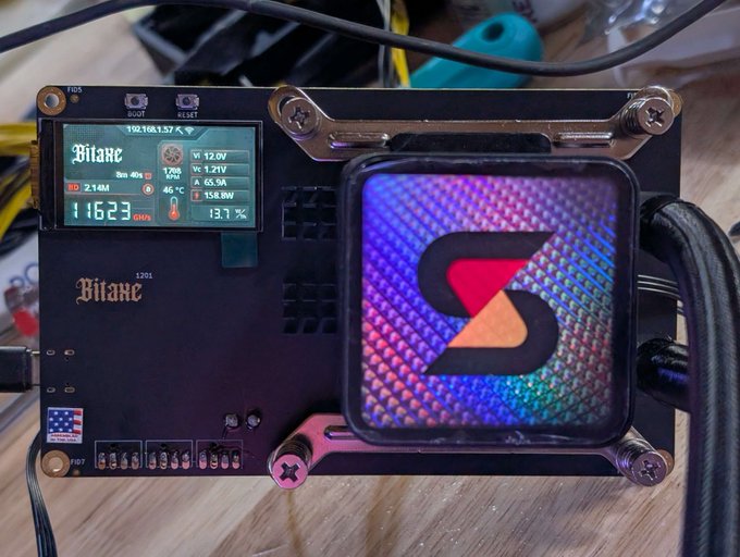 A Bitaxe Naja Duo (model 3201) bitcoin miner is shown running at 116.23 GH/s with 65.5A current draw, displaying a Bitaxe-branded LCD screen with stats including 12.0V, 1.21V voltage, 45°C temperature, and 158.8W power consumption, alongside a square display showing a red and orange "S" logo. The image aligns with the post by illustrating the miner's impressive hashrate output and the addition of a Hydro AIO cooling solution for optimal quietness and efficiency.