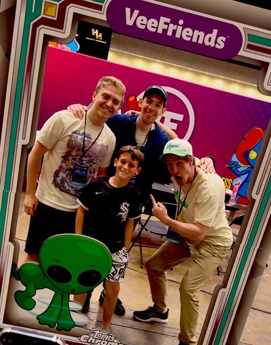 Four people posing for a photo at a 'VeeFriends' booth, with a green alien character graphic in the foreground.
