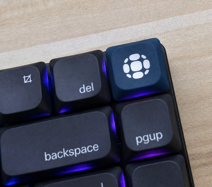 Close-up photo of a mechanical keyboard's upper right corner, highlighting a custom keycap with a white sphere-pattern logo next to the 'backspace' and 'pgup' keys.