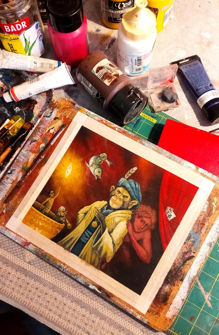 An artist's workspace shows a painted portrait of a green-skinned figure with a blue turban holding a smaller red figure, surrounded by paint tubes, jars, and a cutting mat.
