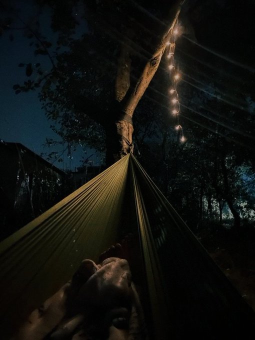 The image shows a person lying down in a hammock under a tree at night with string lights above them. The scene appears to be peaceful, as if someone is taking a break from their daily routine or enjoying some leisure time outdoors. The presence of the string lights adds an element of relaxation and ambiance to the setting.