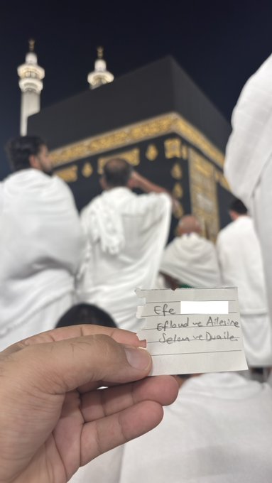 A hand holds a handwritten note reading "Efe, Efclwd-e Atleice, Selen-e Dualer" in the foreground, with pilgrims in ihram gathered around the Kaaba at Masjid al-Haram in Mecca visible in the background, tying the post's gratitude to the sacred setting of worship.