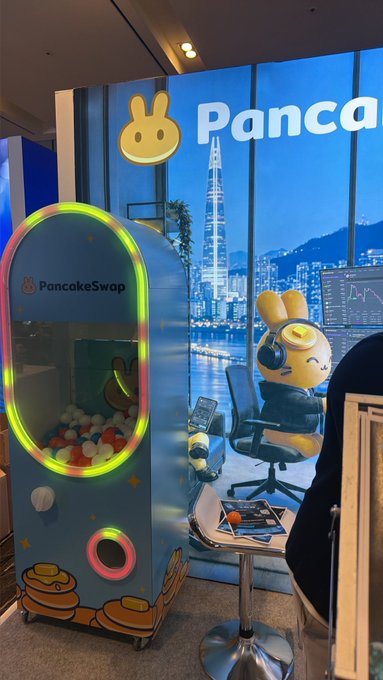 A PancakeSwap branded claw machine filled with colorful balls, situated next to a display featuring a bunny character wearing headphones.