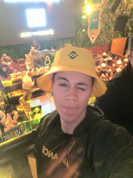 A young person wearing a yellow bucket hat poses for a selfie at what appears to be the Binance Brasil 9th anniversary event, with the Binance logo visible in the background and a yellow "Binance" lanyard around their neck. The lively venue setting supports the caption's description of an exciting celebration with great atmosphere.