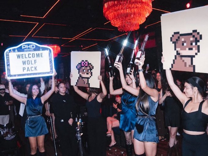 A group of women in dark outfits celebrate at a nightclub, holding up an illuminated sign reading 'PHD WELCOME WOLF PACK' alongside pixel-art signs of a character and bottles of alcohol.