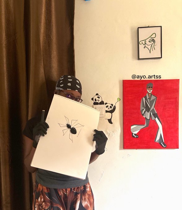 An artist wearing a black bandana and gloves holds up a sketchbook featuring a detailed black widow spider drawing, with the handle "@ayo.artss" visible near a framed line-art piece and a vibrant red painting of a suited figure on the wall behind. The image aligns with the caption introducing Ayo as a traditional artist, showcasing their diverse artistic style ahead of the @ant_clubz NFT collection launch.