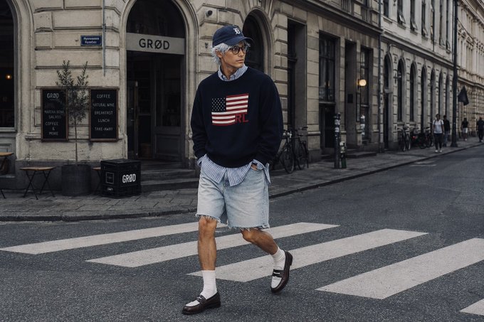 A stylish older man with gray hair and sunglasses crossing a street in a navy "Polo RL" American flag sweater, striped shirt, denim shorts, white socks, and loafers, with a "GRØD" cafe in the background.