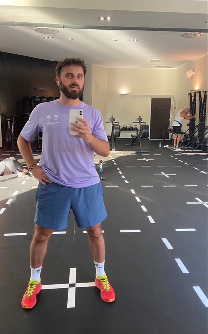 A bearded man takes a mirror selfie at the gym wearing a purple 'Arc'teryx' t-shirt, blue shorts, and bright neon sneakers on a marked training floor.