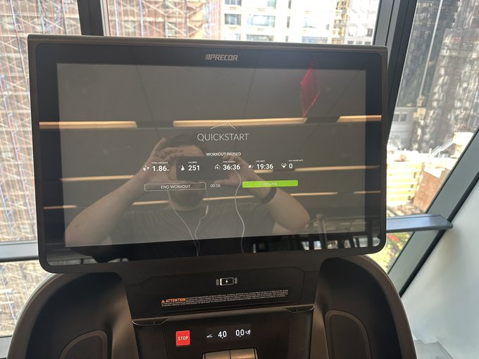 A Precor exercise machine's display screen showing a 'QUICKSTART WORKOUT IN PROGRESS' interface with metrics like distance (1.86) and time (19:36), with the user's reflection visible in the screen