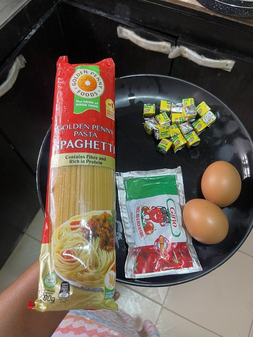 A hand holds a 500g pack of Golden Penny Spaghetti pasta over a black plate containing two brown eggs, a sachet of Gino tomato paste, and several wrapped bouillon cubes. The caption "This dinner go too sweet" likely refers to the small yellow candy-like wrappers among the ingredients, which actually appear to be seasoning cubes rather than sweets.