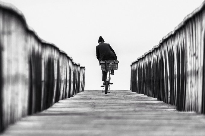 A black and white photograph showing a person riding a bicycle down a wooden boardwalk or pier, moving away from the camera.