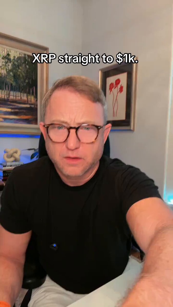 The image features a man wearing glasses sitting at a desk with his arms crossed, looking directly into the camera. The background of the image shows a painting hanging on the wall behind him. There are also two other people visible in the image, one located near the top left corner and another person closer to the center-right side of the frame.