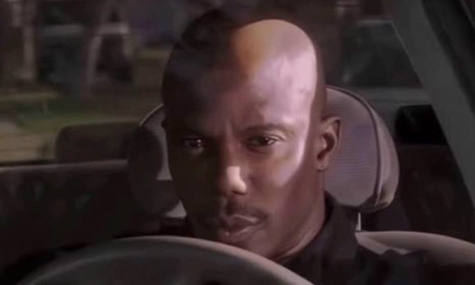 A close-up of Dave Chappelle sitting in the driver's seat of a car with a serious expression.
