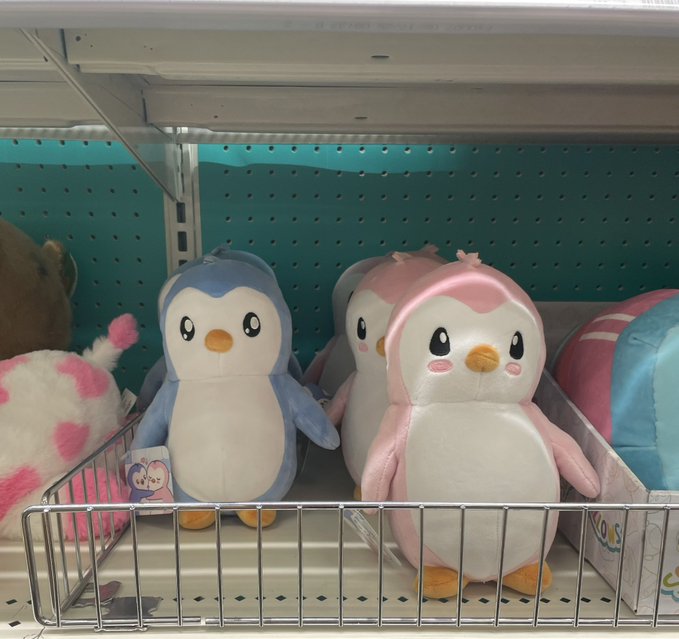 Plush toys of Pengu (the blue penguin) and Polly (the pink penguin) are displayed on a store shelf, representing the Pudgy Penguins NFT brand's retail expansion that drives visibility for the $PENGU token.