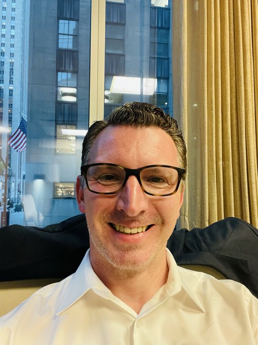 A selfie of a smiling man with short hair and glasses wearing a white shirt, with a window showing city buildings and an American flag behind him.