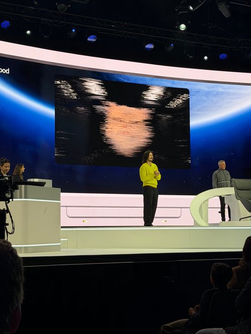 A woman in a yellow sweater speaks on stage at a Robinhood event, standing before a large screen displaying an abstract orange and black image, with a man in a gray sweater visible to her right. The partially visible "ood" text on the upper left of the screen and the "Robinhood" branding align with the context of CEO Vlad Tenev discussing his mission to democratize finance.