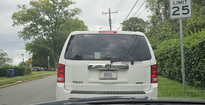 Rear view of a white Honda Pilot SUV with a personalized license plate reading "HODL," stopped on a road next to a 25 mph speed limit sign.