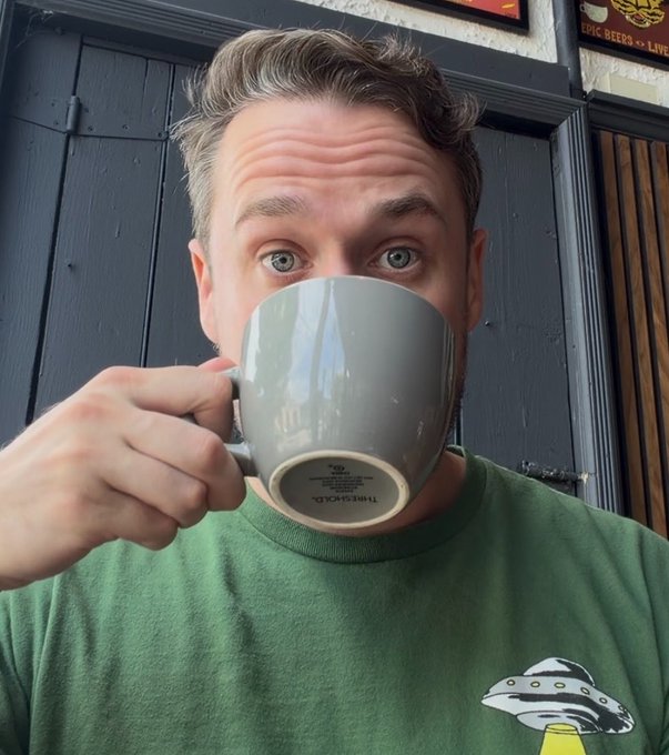A man in a green t-shirt takes a selfie while drinking from a large gray mug, looking directly at the camera with wide eyes in what appears to be a pub setting.