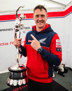 The image features a man wearing a red hoodie, holding a trophy with both hands while smiling at the camera. The trophy appears to be made of metal or silver material, possibly a trophy from a racing event. The background shows a white tent, suggesting that this moment might have taken place in an outdoor setting like a race track or stadium. The post on which the image is displayed reads "BCH & Deano FTW! @deanharrisonTT". This indicates that the man holding the trophy could be a professional racer or athlete who has achieved success in their sport, and they are sharing this moment with others through social media platforms.