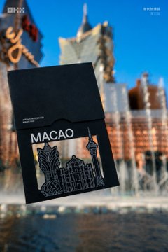 A promotional image showing a dark-colored booklet or passport labeled 'MACAO' with a silver line-art illustration of the Macau skyline, set against a blurred background of Macau's architecture and waterfront.