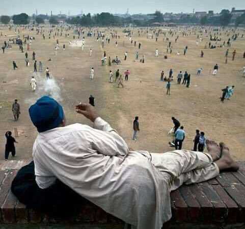 A man in traditional attire reclines on a brick ledge, casually smoking while observing a large crowd playing cricket on an expansive dusty field below. The image humorously captures a sense of leisurely detachment from the chaotic activity, mirroring the post's sentiment about being unbothered while others are caught up in the "Robinhood chain."