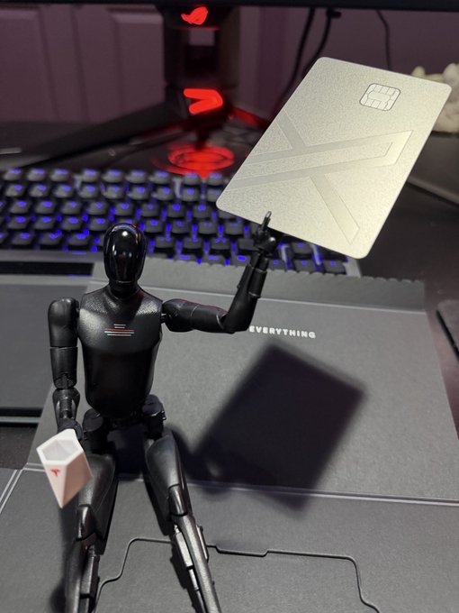 A black action figure sits on a desk mat in front of a keyboard, holding up a silver card embossed with a Union Jack design.
