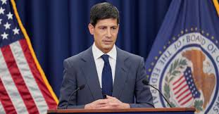 A man in a dark suit and blue tie speaks at a podium with a Federal Reserve Board seal and American flag visible behind him.