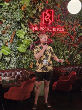 A man stands in front of a neon sign reading 'THE BROKERS BAR,' holding a microphone as if performing karaoke, with a wall of greenery behind him.