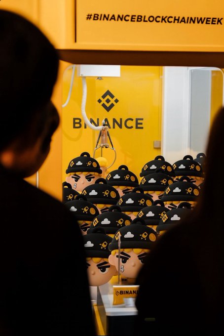A Binance claw machine branded with "#BINANCEBLOCKCHAINWEEK" displays plush Binance mascot toys, seen past the silhouettes of attendees. The image showcases a crypto-themed promotional event, though it doesn't directly connect to the $PALU token or Chinese Valentine's Day narrative mentioned in the post.