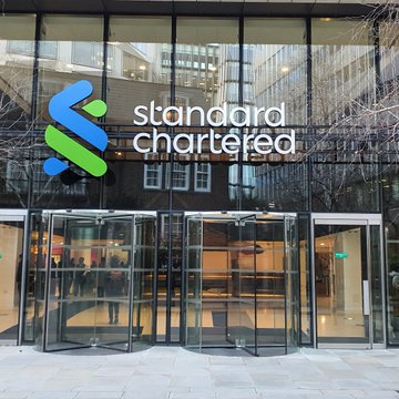 The exterior entrance of a Standard Chartered bank building features the company's blue and green logo on a glass facade above a revolving door.