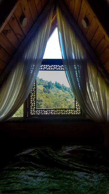 A view from inside a wooden cabin with a triangular window framed by curtains, looking out onto a green hillside.